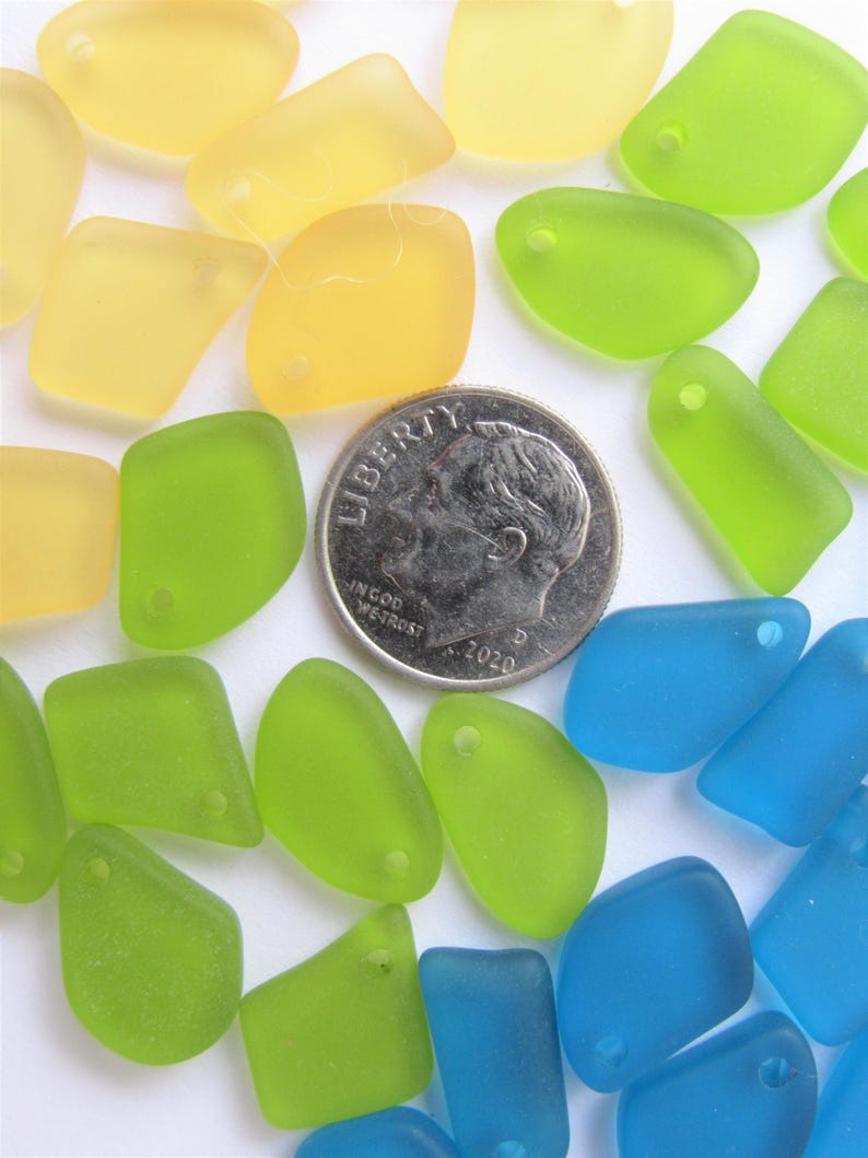 May include: A collection of translucent, frosted glass pieces in yellow, lime green, and blue, each featuring a small pre-drilled hole for jewelry making. A silver-colored United States dime is positioned among the pieces to provide a scale reference.