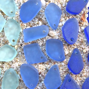 May include: A collection of frosted sea glass pieces in various shades of blue, ranging from pale aquamarine to deep cobalt. Each piece features a smooth, weathered texture and a single pre-drilled hole, making them ready for jewelry making.