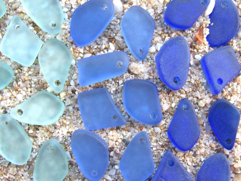 May include: A collection of frosted sea glass pieces in various shades of blue, ranging from pale aquamarine to deep cobalt. Each piece features a smooth, weathered texture and a single pre-drilled hole, making them ready for jewelry making.