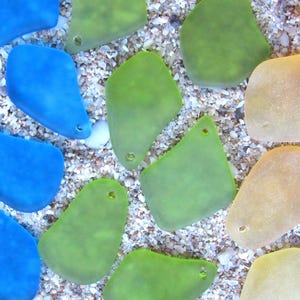 May include: An overhead shot of sea glass pieces in shades of blue, green, and tan. The pieces are irregularly shaped with smooth edges and small holes. They are scattered on a bed of small, light-colored pebbles and sand.