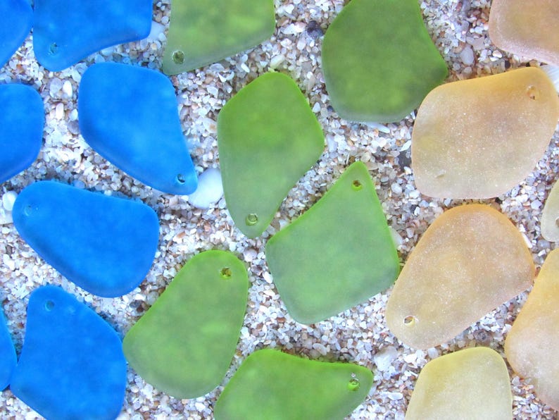 May include: An overhead shot of sea glass pieces in shades of blue, green, and tan. The pieces are irregularly shaped with smooth edges and small holes. They are scattered on a bed of small, light-colored pebbles and sand.