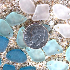 May include: A collection of frosted, translucent sea glass pieces in shades of white, light blue, and dark blue, each featuring a single pre-drilled hole. A silver-colored United States dime is placed in the center for scale reference.