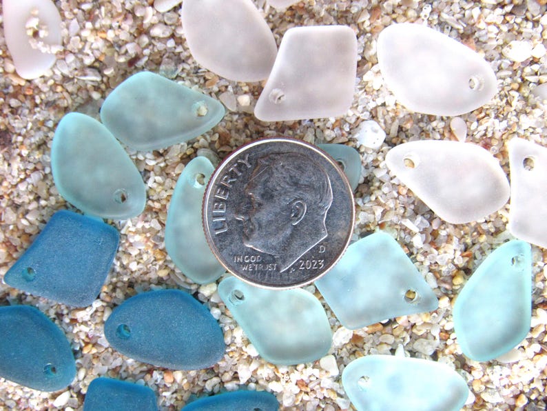 May include: A collection of frosted, translucent sea glass pieces in shades of white, light blue, and dark blue, each featuring a single pre-drilled hole. A silver-colored United States dime is placed in the center for scale reference.