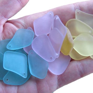 May include: A handful of sea glass pieces in various pastel colors: light blue, lavender, and yellow. Each piece has a smooth, frosted appearance and a small hole for stringing. The sea glass is in a variety of shapes, including rounded and rectangular.
