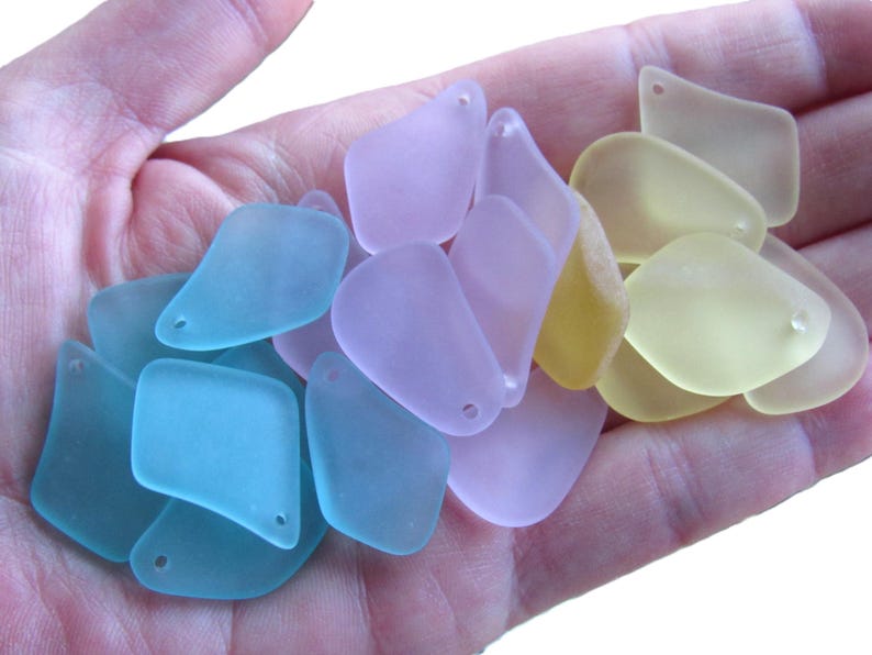 May include: A handful of sea glass pieces in various pastel colors: light blue, lavender, and yellow. Each piece has a smooth, frosted appearance and a small hole for stringing. The sea glass is in a variety of shapes, including rounded and rectangular.