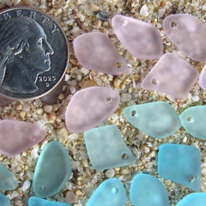 May include: A collection of small, frosted, irregular-shaped glass beads in shades of soft pink and light blue, each featuring a single drilled hole. A United States quarter coin is included for scale, measuring approximately 0.955 inches in diameter.