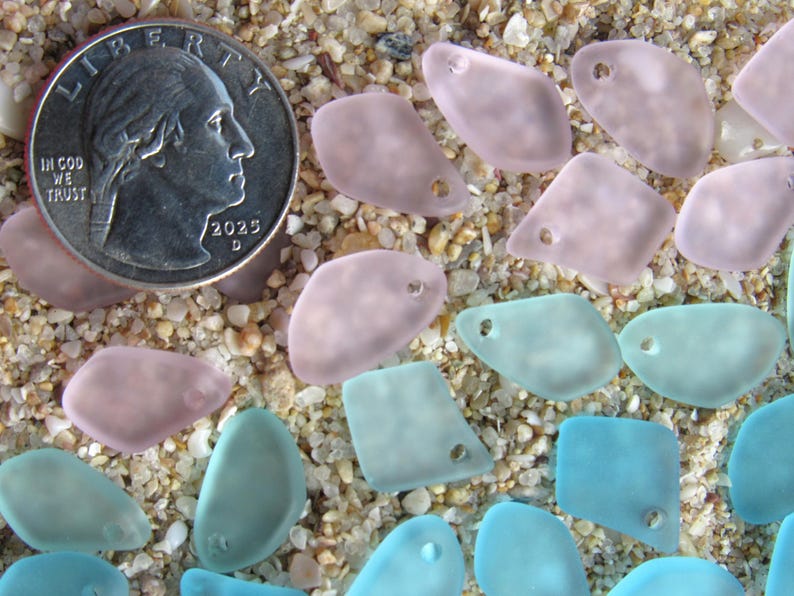May include: A collection of small, frosted, irregular-shaped glass beads in shades of soft pink and light blue, each featuring a single drilled hole. A United States quarter coin is included for scale, measuring approximately 0.955 inches in diameter.
