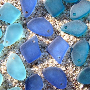 May include: A collection of frosted sea glass pieces in various shades of blue, ranging from light aqua to deep cobalt. Each piece features a smooth, weathered texture and a single pre-drilled hole, making them ready for use as jewelry components.