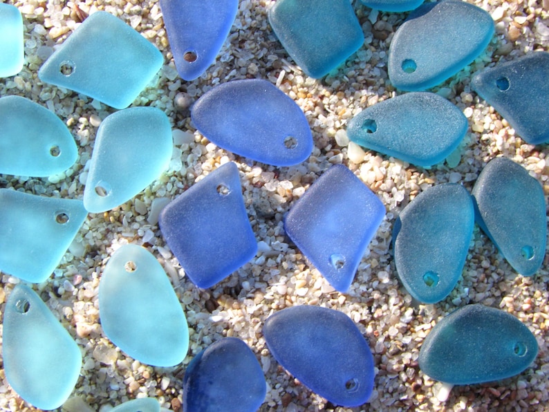 May include: A collection of frosted sea glass pieces in various shades of blue, ranging from light aqua to deep cobalt. Each piece features a smooth, weathered texture and a single pre-drilled hole, making them ready for use as jewelry components.