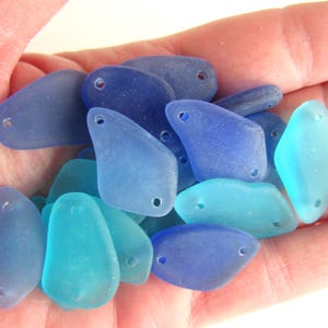 May include: A collection of sea glass charms in shades of blue and turquoise. Each charm has two holes for attaching to jewelry or other crafts.
