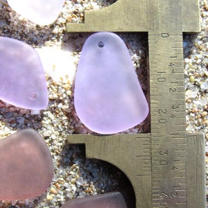 May include: Close-up of sea glass pieces in shades of lavender, pink, and clear, with a brass caliper measuring one piece. The sea glass has a smooth, frosted appearance. The caliper shows a measurement of approximately 1.5 inches.