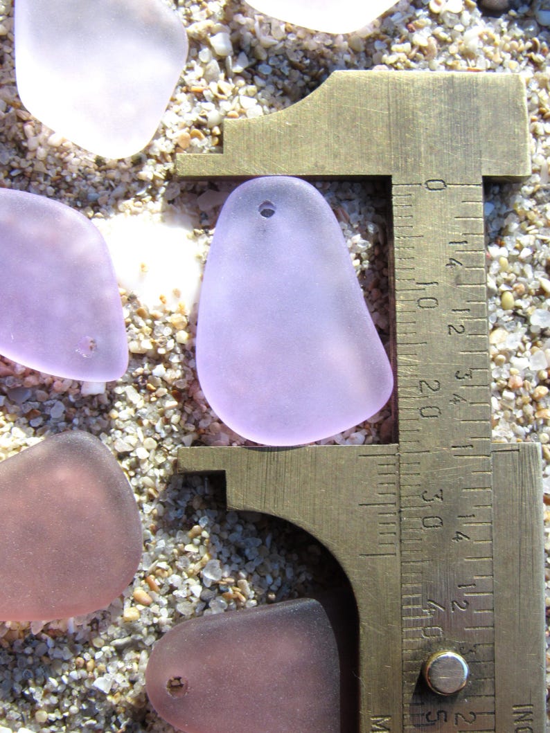 May include: Close-up of sea glass pieces in shades of lavender, pink, and clear, with a brass caliper measuring one piece. The sea glass has a smooth, frosted appearance. The caliper shows a measurement of approximately 1.5 inches.