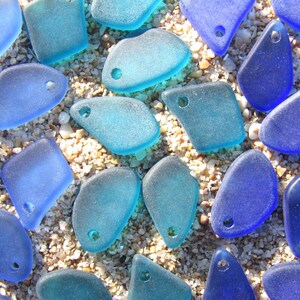 May include: A collection of assorted sea glass pieces in various shades of blue and turquoise. Each piece features a smooth, frosted texture and a single pre-drilled hole, making them ready for use as individual jewelry components or charms.