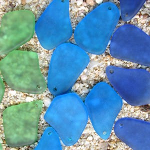 May include: An overhead shot of sea glass pieces in shades of blue and green. The pieces are irregularly shaped with a frosted appearance and small holes. They are scattered on a bed of sand and small pebbles.