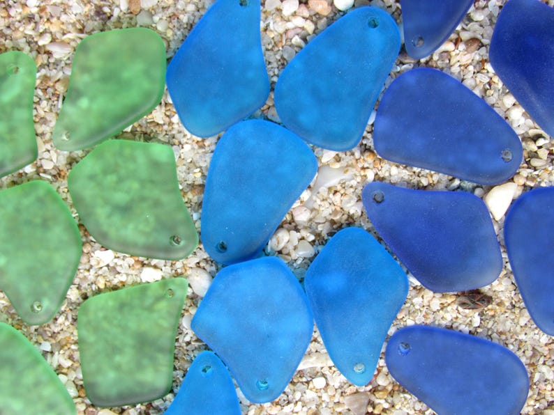 May include: An overhead shot of sea glass pieces in shades of blue and green. The pieces are irregularly shaped with a frosted appearance and small holes. They are scattered on a bed of sand and small pebbles.