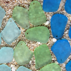 May include: An overhead shot of sea glass pieces in shades of blue and green, scattered on a sandy surface. The pieces have a frosted appearance and are irregularly shaped, with small holes drilled at the top. The background is a mix of sand and small pebbles.