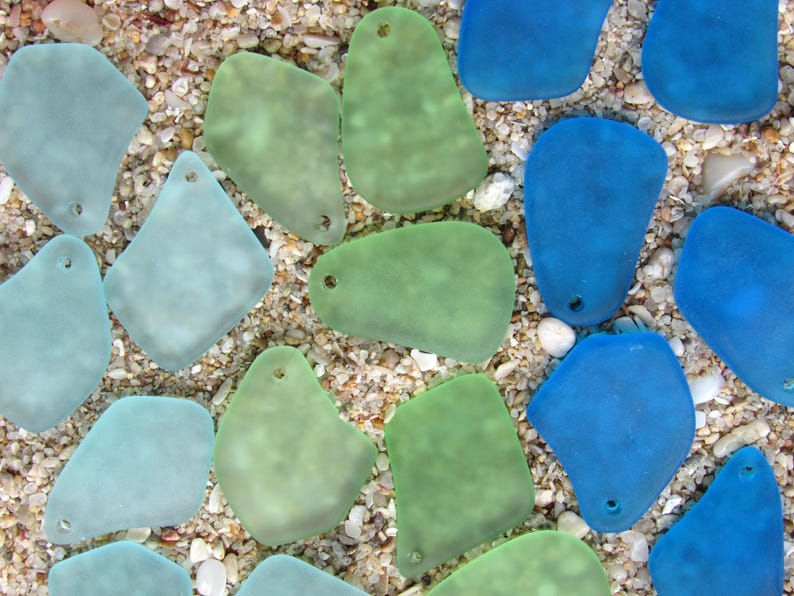 May include: An overhead shot of sea glass pieces in shades of blue and green, scattered on a sandy surface. The pieces have a frosted appearance and are irregularly shaped, with small holes drilled at the top. The background is a mix of sand and small pebbles.
