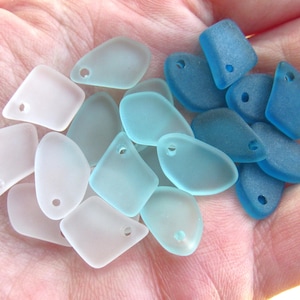 May include: A collection of sea glass beads in shades of white, light blue, and blue. The beads have a smooth, frosted appearance and are drilled with a small hole for stringing. The shapes vary from rounded to geometric.