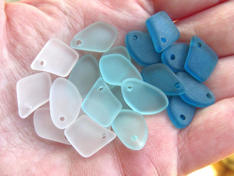 May include: A collection of frosted glass pendants in shades of pale pink, light blue, and deep ocean blue. Each piece features a smooth, rounded, irregular shape with a single pre-drilled hole near the top edge for stringing or jewelry making.