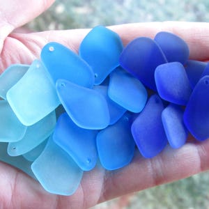May include: A collection of sea glass pieces in varying shades of blue, from light turquoise to deep indigo. Each piece has a smooth, frosted texture and a small hole for stringing. The pieces are held in a hand.