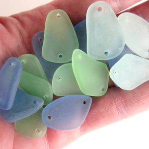 May include: A collection of sea glass pieces in various shades of blue, green, and white. Each piece has a hole drilled through it, suggesting they are intended for jewelry making or crafting.