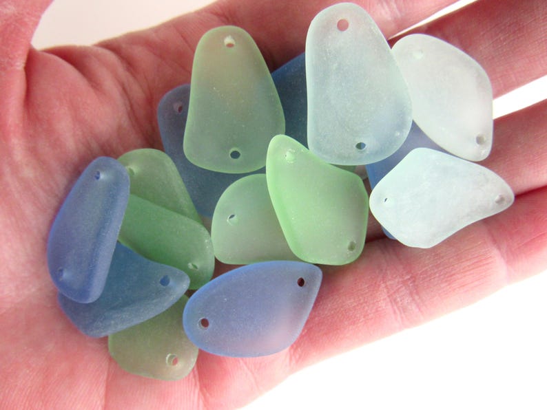 May include: A collection of sea glass pieces in various shades of blue, green, and white. Each piece has a hole drilled through it, suggesting they are intended for jewelry making or crafting.