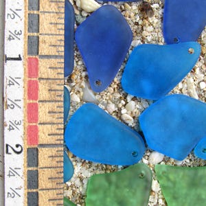 May include: Close-up of sea glass pieces in shades of blue and green, with a wooden ruler showing measurements in inches. The sea glass is scattered on a bed of sand and small pebbles. Each piece has a small hole.
