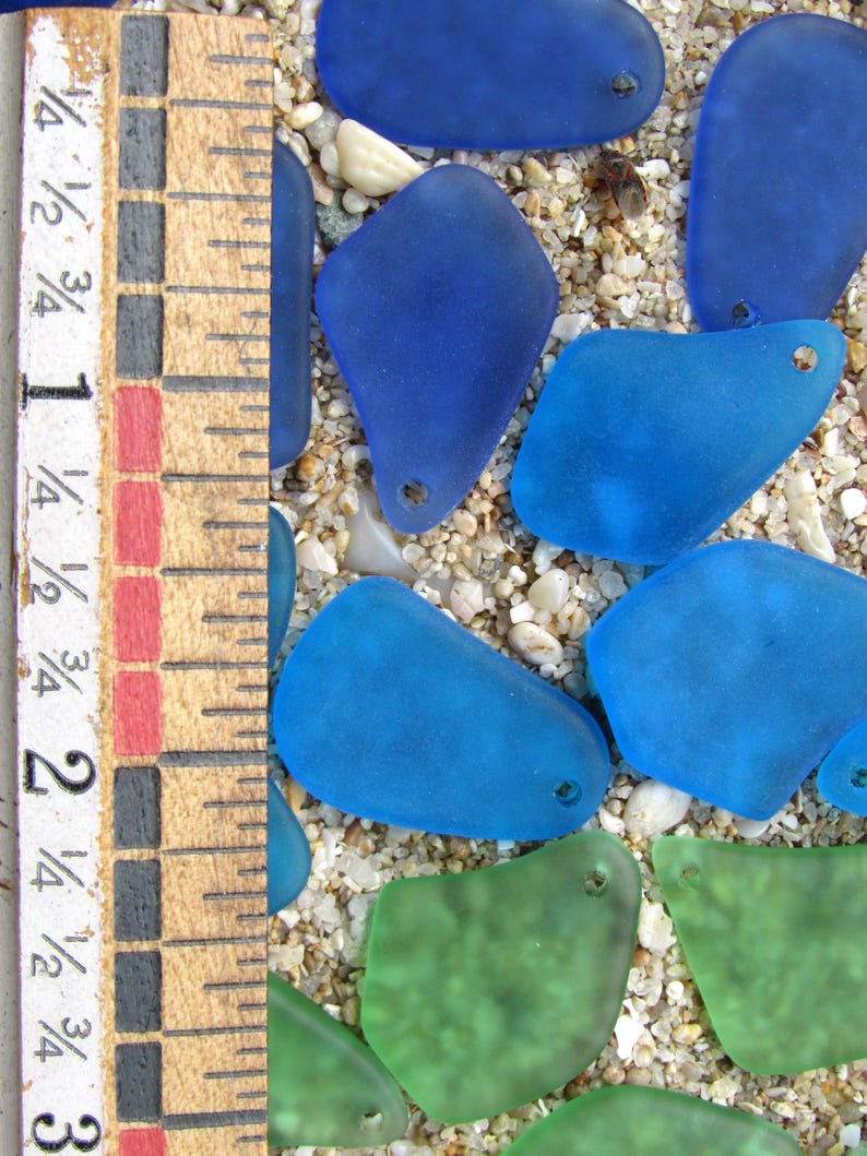 May include: Close-up of sea glass pieces in shades of blue and green, with a wooden ruler showing measurements in inches. The sea glass is scattered on a bed of sand and small pebbles. Each piece has a small hole.