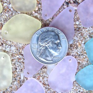 May include: A quarter coin rests among sea glass pieces in shades of yellow, lavender, and blue. The sea glass has a smooth, frosted appearance and is scattered on a sandy surface. The coin is dated 2022.