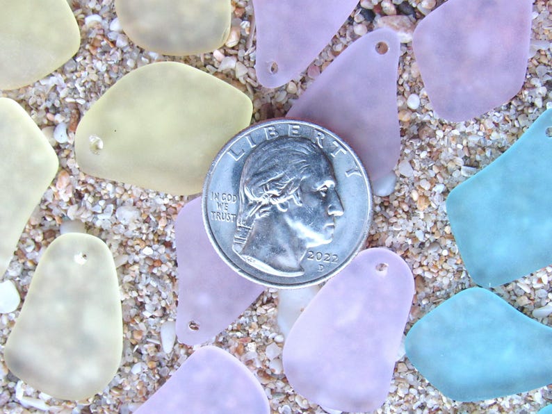 May include: A quarter coin rests among sea glass pieces in shades of yellow, lavender, and blue. The sea glass has a smooth, frosted appearance and is scattered on a sandy surface. The coin is dated 2022.