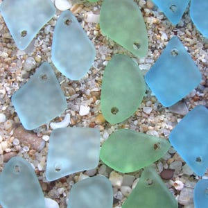 May include: A collection of frosted, translucent sea glass pendants in various shades of light blue, aqua, and seafoam green. Each piece features a smooth, weathered texture and a single pre-drilled hole at the top for stringing or jewelry making.