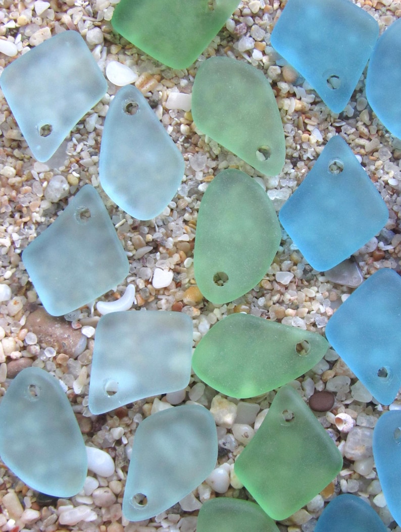 May include: A collection of frosted, translucent sea glass pendants in various shades of light blue, aqua, and seafoam green. Each piece features a smooth, weathered texture and a single pre-drilled hole at the top for stringing or jewelry making.