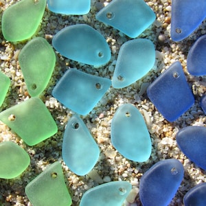 May include: A collection of smooth, frosted sea glass pieces in various shades of green, light blue, and dark blue. Each piece features a single pre-drilled hole near the top edge, creating a uniform appearance for use as jewelry components.