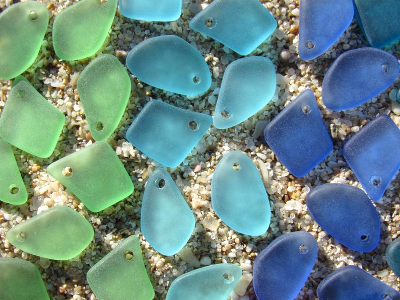 May include: A collection of smooth, frosted sea glass pieces in various shades of green, light blue, and dark blue. Each piece features a single pre-drilled hole near the top edge, creating a uniform appearance for use as jewelry components.