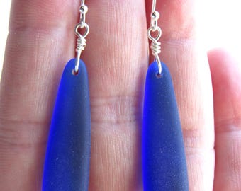 Könnte beinhalten: Ein Paar Ohrringe mit tropfenförmigen Anhängern aus tiefblauem Meeresglas. Die Anhänger hängen an silberfarbenen Haken mit Drahtdetails. Das Design ist schlicht und elegant.