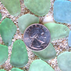 May include: An overhead shot of sea glass pieces in shades of green and blue, scattered on a sandy surface. A U.S. quarter coin is centered among the glass, providing a size reference. The sea glass pieces have a smooth, frosted appearance.