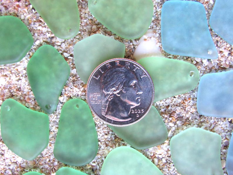 May include: An overhead shot of sea glass pieces in shades of green and blue, scattered on a sandy surface. A U.S. quarter coin is centered among the glass, providing a size reference. The sea glass pieces have a smooth, frosted appearance.