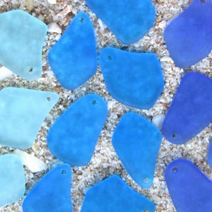 May include: An overhead shot of sea glass pieces in various shades of blue and aqua. The pieces are irregularly shaped with smooth edges and small holes. They are scattered on a bed of sand and small shells.