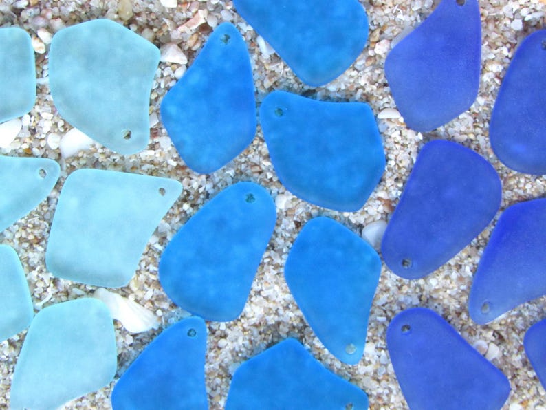 May include: An overhead shot of sea glass pieces in various shades of blue and aqua. The pieces are irregularly shaped with smooth edges and small holes. They are scattered on a bed of sand and small shells.