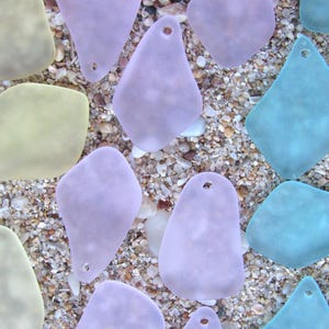 May include: An assortment of sea glass pieces in various shapes and pastel colors, including lavender, teal, and pale yellow. Each piece has a small hole and rests on a bed of sand and small pebbles, creating a beachy aesthetic.