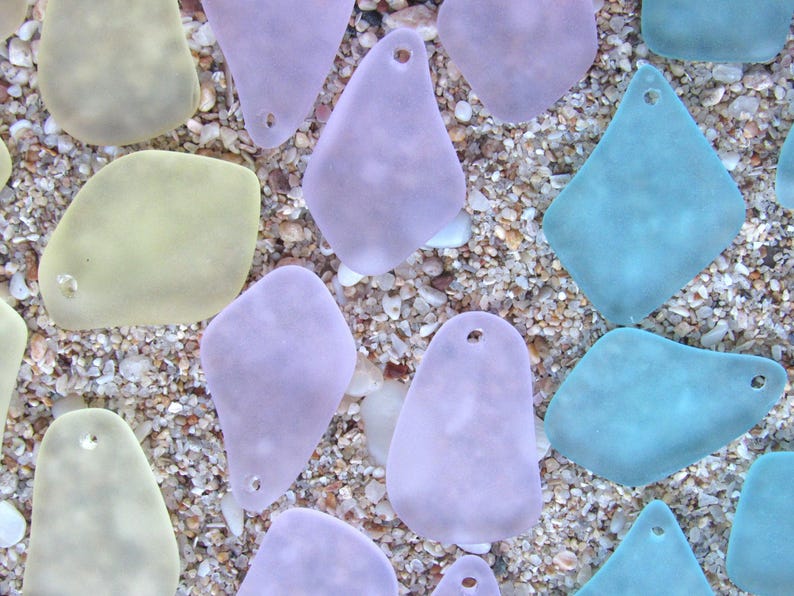 May include: An assortment of sea glass pieces in various shapes and pastel colors, including lavender, teal, and pale yellow. Each piece has a small hole and rests on a bed of sand and small pebbles, creating a beachy aesthetic.