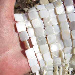 May include: A strand of white square-shaped mother of pearl beads. The beads are smooth and have a slight iridescent sheen.