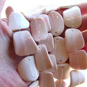 May include: A strand of smooth, off-white, irregularly shaped beads with a glossy finish. Each bead features a small, centered hole for stringing. The beads are arranged in a cluster, showcasing their varied, organic, and rounded rectangular forms.