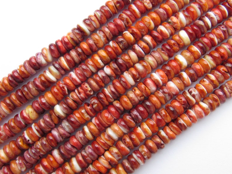 Peut inclure: Gros plan sur des brins de perles en coquillage d'hu&icirc;tre &eacute;pineuse. Les perles sont un m&eacute;lange de couleurs brun rouge&acirc;tre, orange et blanc. Les perles sont petites, rondes et de forme l&eacute;g&egrave;rement irr&eacute;guli&egrave;re. Les perles sont enfil&eacute;es en rang&eacute;e.