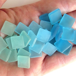 Könnte beinhalten: Eine Sammlung quadratischer Seeglasfliesen in verschiedenen Blau- und Aquatönen. Die Fliesen werden in einer Hand gehalten und zeigen ihre matte Textur und unregelmäßige Formen. Ideal für Mosaike oder Bastelarbeiten.