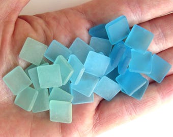 May include: A collection of square, sea glass tiles in varying shades of blue and aqua. The tiles are held in a hand, showcasing their frosted texture and irregular shapes. Ideal for mosaics or crafts.