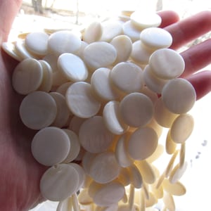 May include: A pile of white round mother of pearl beads. The beads are smooth and have a slight iridescent sheen.