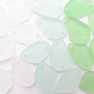 May include: A collection of smooth, frosted glass sea glass pieces in various shades of green and white. The pieces are shaped like teardrops and have two holes drilled through them.