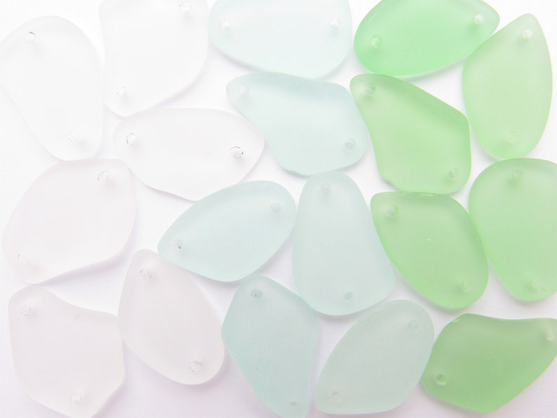 May include: A collection of smooth, frosted glass sea glass pieces in various shades of green and white. The pieces are shaped like teardrops and have two holes drilled through them.