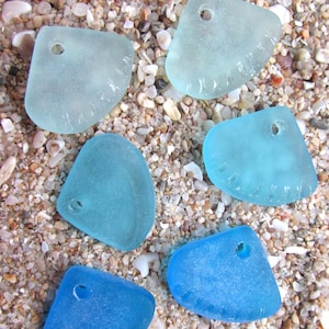 May include: Six pieces of sea glass in varying shades of blue and aqua, each with a drilled hole for crafting. The sea glass is arranged on a bed of sand and small pebbles, creating a natural, beachy aesthetic.